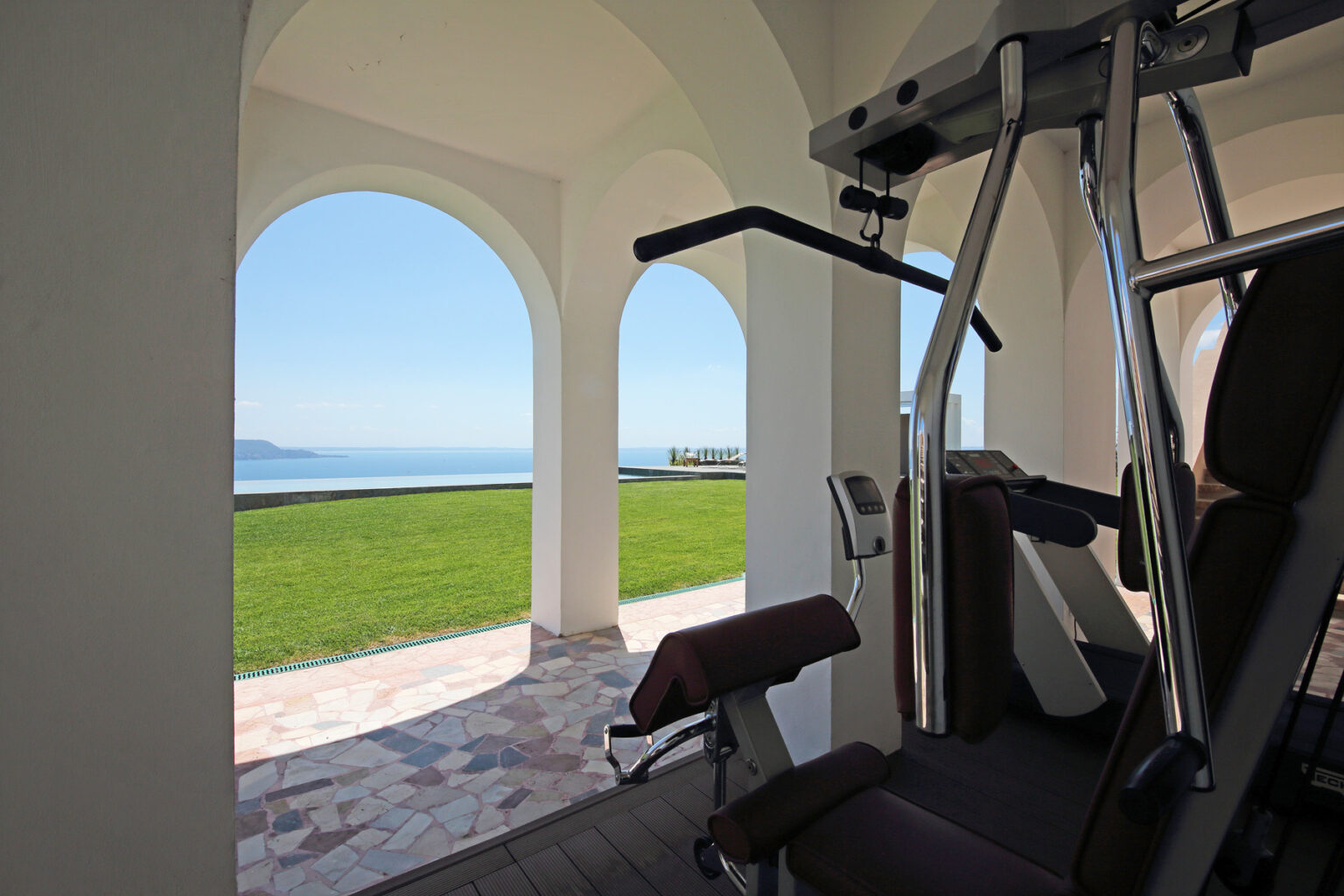 Home gym with a view of the ocean, featuring exercise equipment framed by elegant arches and lush green grass outside. | Only on SopranoVillas