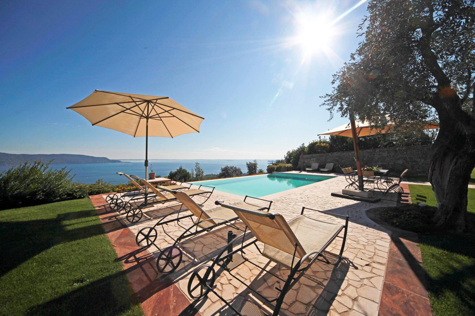 Luxury villa with a serene poolside view overlooking the ocean, featuring sun loungers and umbrellas under bright sunlight, surrounded by lush greenery. Perfect for a relaxing getaway. | Only on SopranoVillas