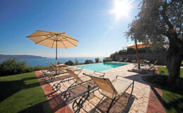 Luxury villa with a serene poolside view overlooking the ocean, featuring sun loungers and umbrellas under bright sunlight, surrounded by lush greenery. Perfect for a relaxing getaway.