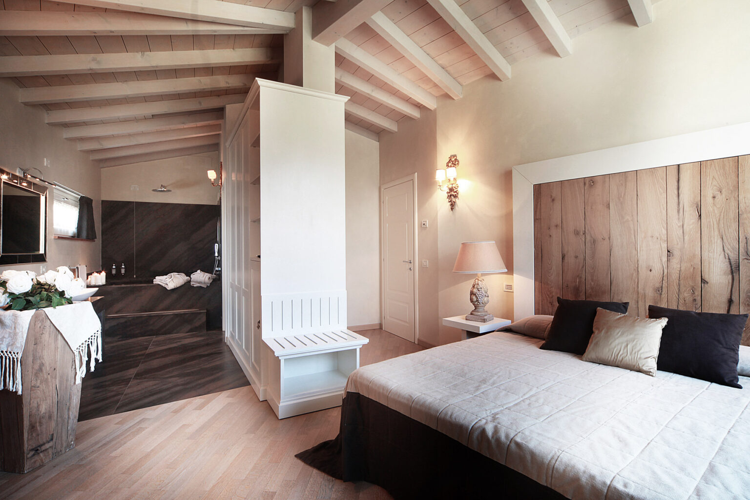 Modern bedroom interior featuring a queen-sized bed with decorative pillows, wooden accents, and a stylish ensuite bathroom. The room has high ceilings with exposed beams, soft lighting, and a cozy atmosphere, ideal for relaxation. | Only on SopranoVillas