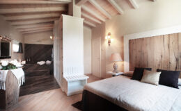 Modern bedroom interior featuring a queen-sized bed with decorative pillows, wooden accents, and a stylish ensuite bathroom. The room has high ceilings with exposed beams, soft lighting, and a cozy atmosphere, ideal for relaxation.