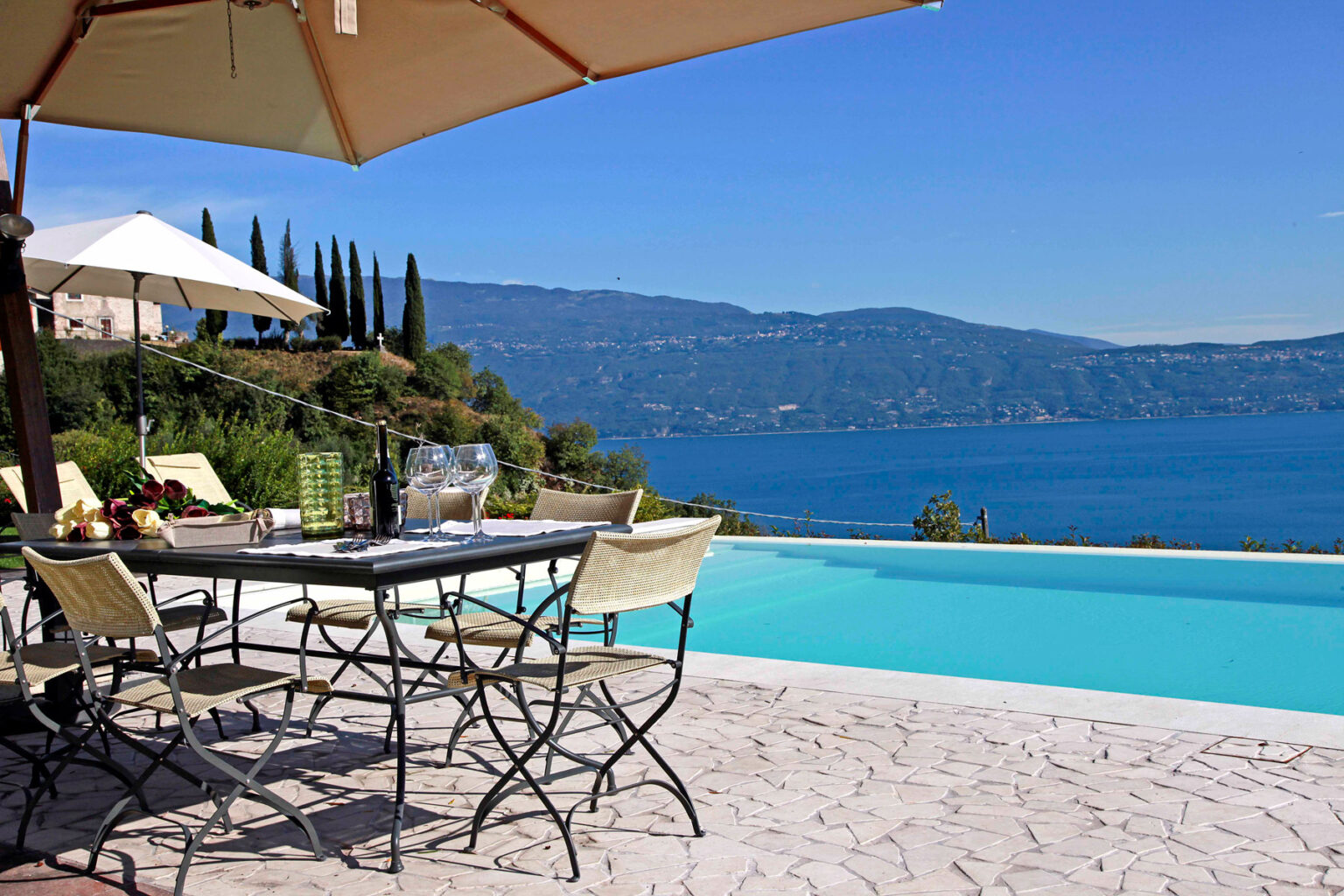 Outdoor dining area by a luxury pool overlooking a serene lake, surrounded by lush greenery and distant mountains under a clear blue sky. | Only on SopranoVillas