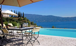 Outdoor dining area by a luxury pool overlooking a serene lake, surrounded by lush greenery and distant mountains under a clear blue sky.