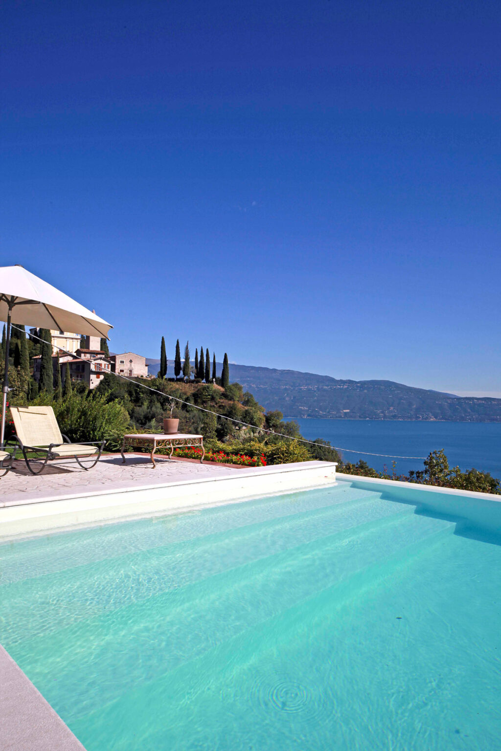 Luxury villa with a swimming pool overlooking a serene lake and mountains, featuring a sun lounger and parasol in a beautifully landscaped garden. | Only on SopranoVillas