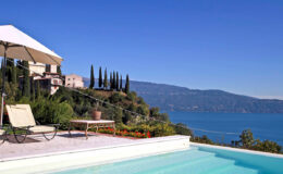 Luxury villa with a swimming pool overlooking a serene lake and mountains, featuring a sun lounger and parasol in a beautifully landscaped garden.