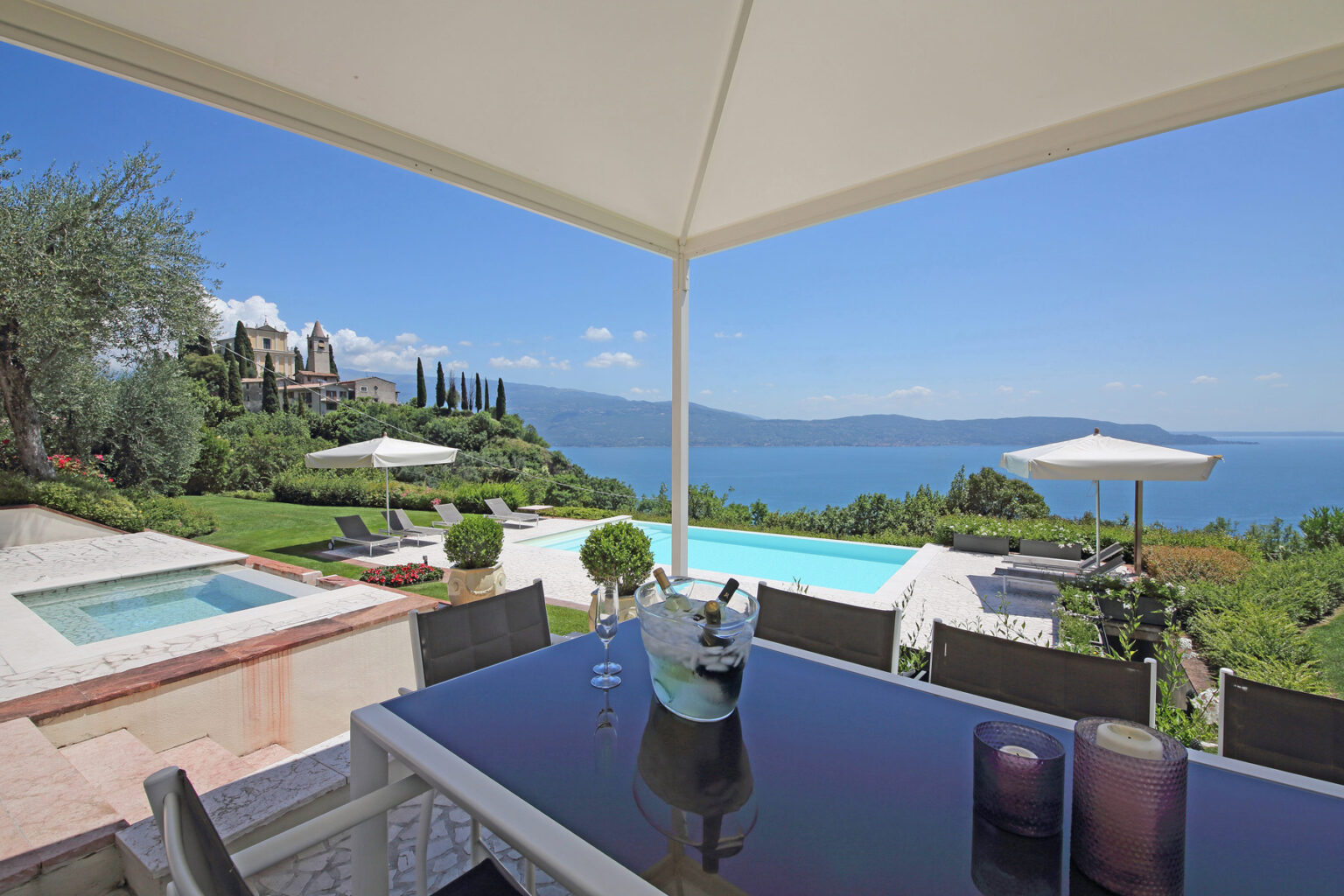 Luxurious villa patio with a stunning view of a lake, featuring a modern dining table, outdoor pool, and manicured gardens under a clear blue sky. | Only on SopranoVillas
