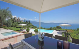 Luxurious villa patio with a stunning view of a lake, featuring a modern dining table, outdoor pool, and manicured gardens under a clear blue sky.