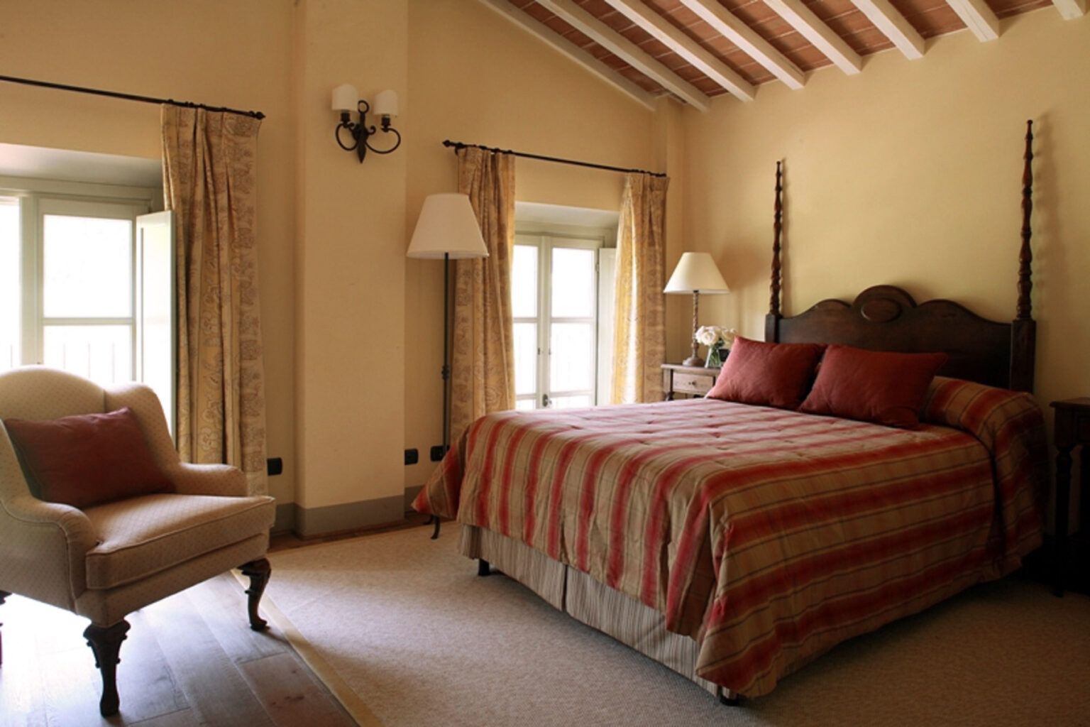 Cozy bedroom featuring a queen-sized bed with a striped red and beige comforter, elegant wooden headboard, and soft lighting from bedside lamps. A comfortable armchair is positioned near the window, enhancing the inviting atmosphere of the room. | Only on SopranoVillas