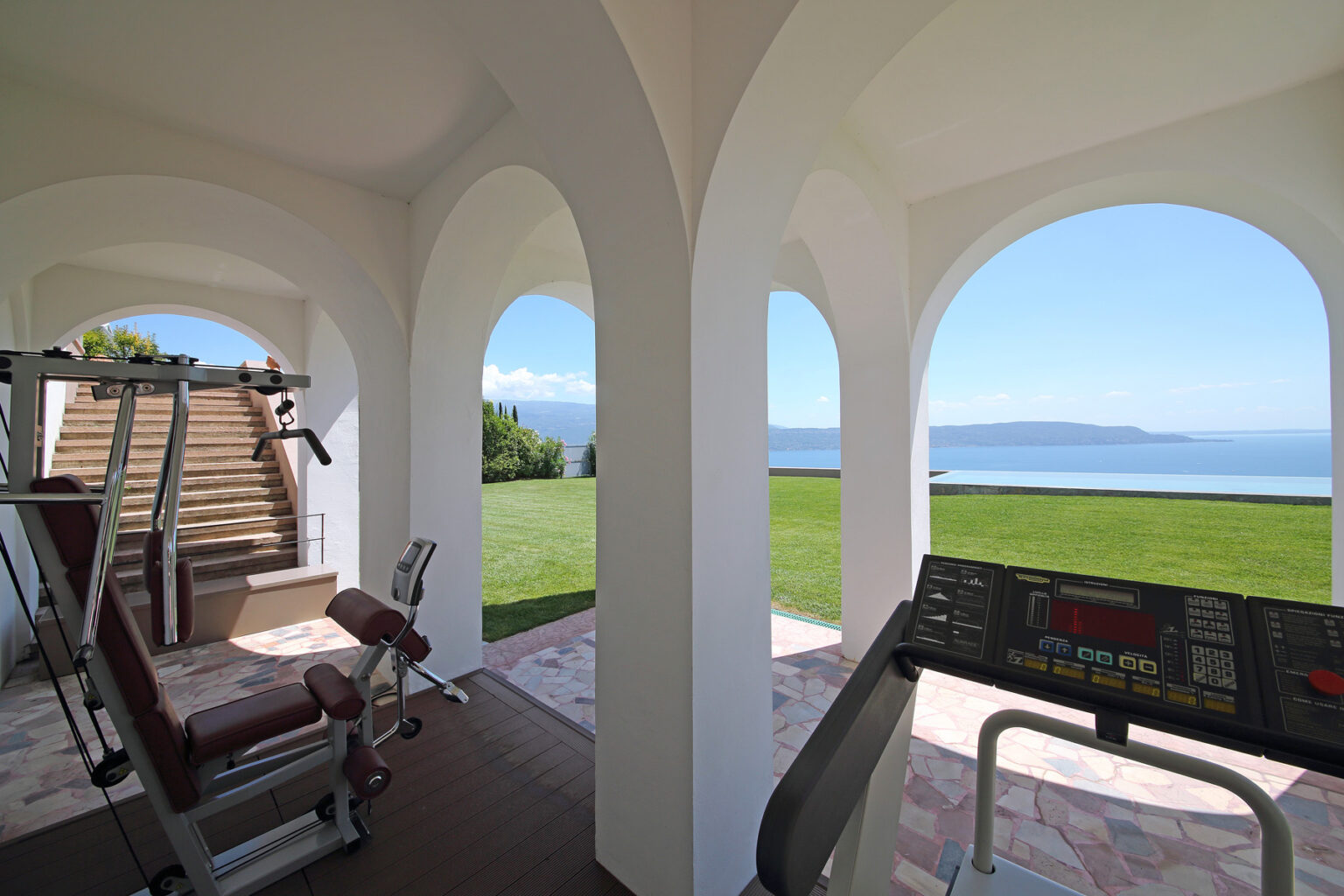 Home gym with panoramic views of a serene landscape, featuring exercise equipment like a treadmill and weight machines, set in a stylish architectural space with arched openings. | Only on SopranoVillas