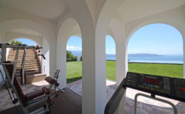 Home gym with panoramic views of a serene landscape, featuring exercise equipment like a treadmill and weight machines, set in a stylish architectural space with arched openings.