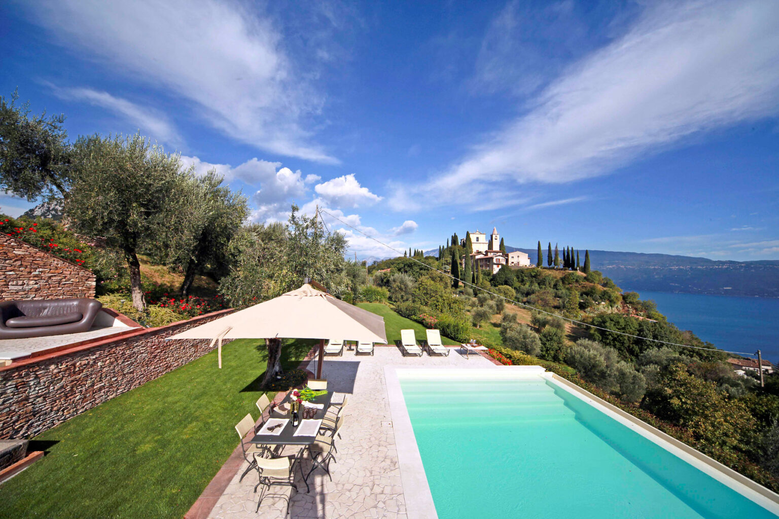 Scenic view of a luxurious villa with a swimming pool, surrounded by olive trees and lush greenery, overlooking a serene lake and distant hills under a blue sky. | Only on SopranoVillas