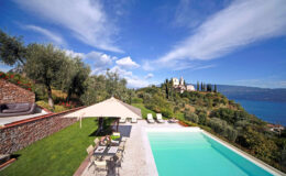 Scenic view of a luxurious villa with a swimming pool, surrounded by olive trees and lush greenery, overlooking a serene lake and distant hills under a blue sky.