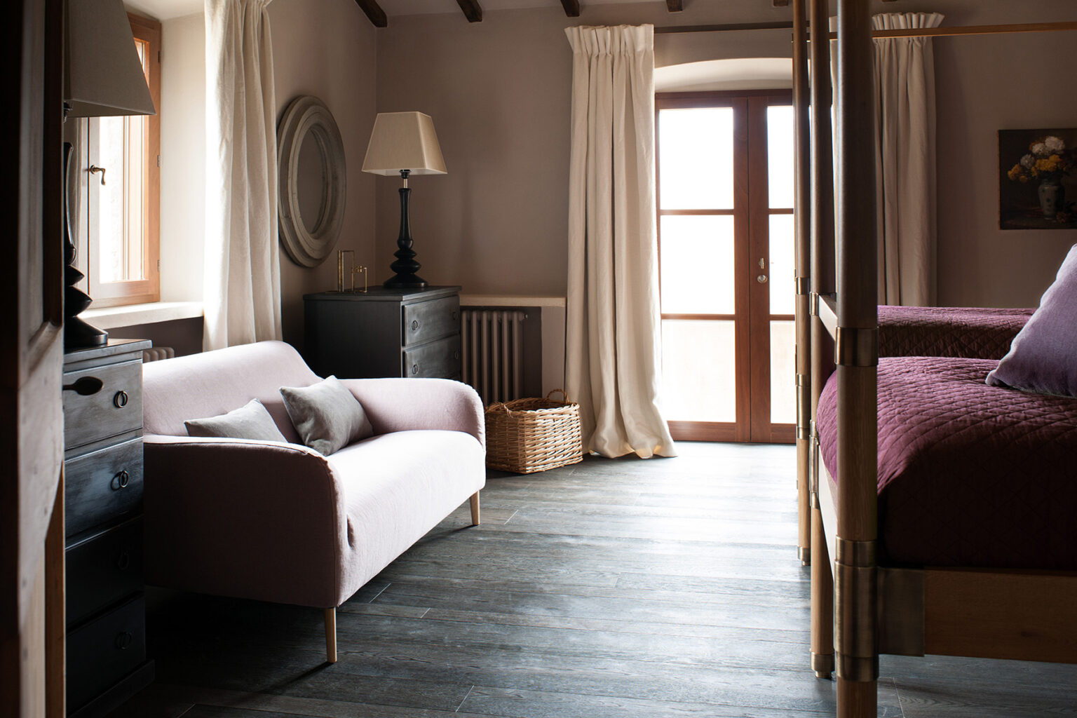 Cozy bedroom interior featuring a pink sofa, a wooden bed with a maroon blanket, and large windows with sheer curtains allowing natural light. The room includes a stylish lamp and a decorative basket, enhancing the warm and inviting atmosphere. | Only on SopranoVillas