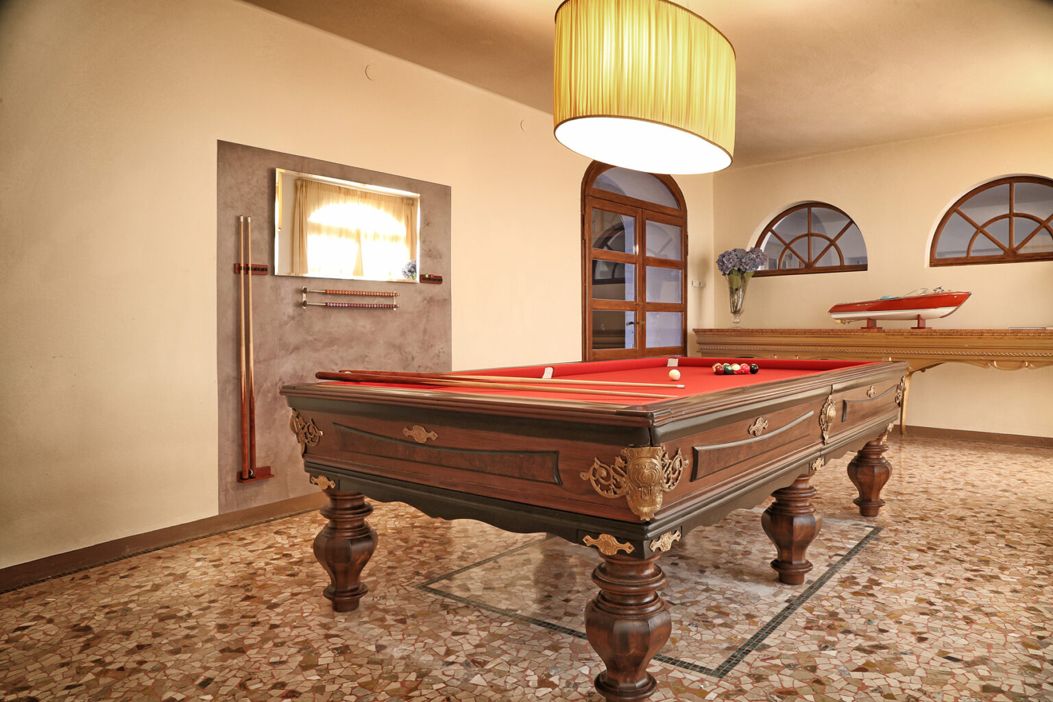 Elegant game room featuring a vintage billiard table with a red felt surface, surrounded by neutral-toned walls and decorative windows. A stylish pendant lamp hangs above, enhancing the inviting atmosphere. | Only on SopranoVillas