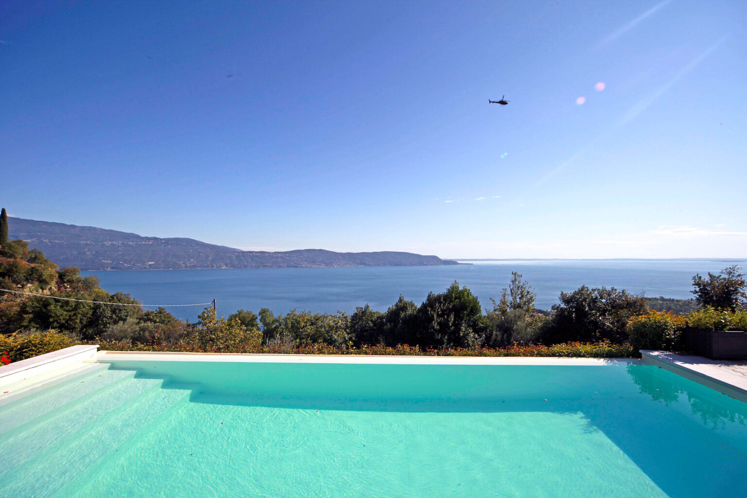 Scenic view of a tranquil swimming pool overlooking a serene lake and distant mountains under a clear blue sky. | Only on SopranoVillas