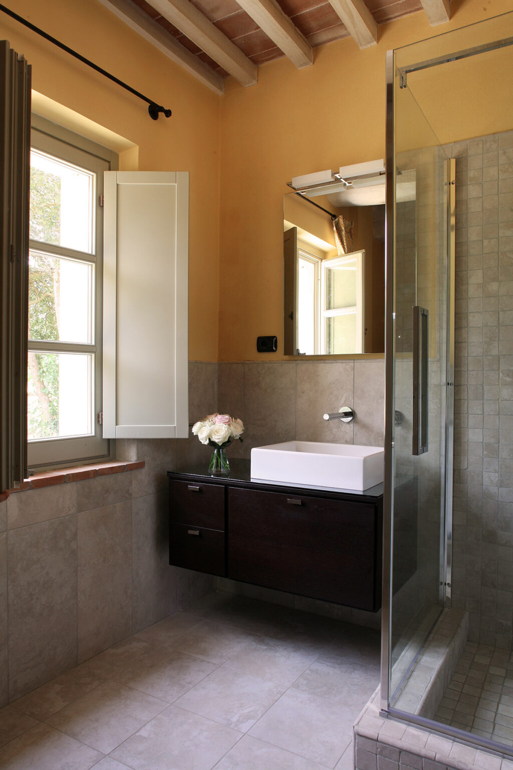 Modern bathroom design featuring a sleek white sink, dark wood cabinetry, and a glass shower. Natural light enters through an open window, complemented by a vase of fresh flowers on the counter. | Only on SopranoVillas