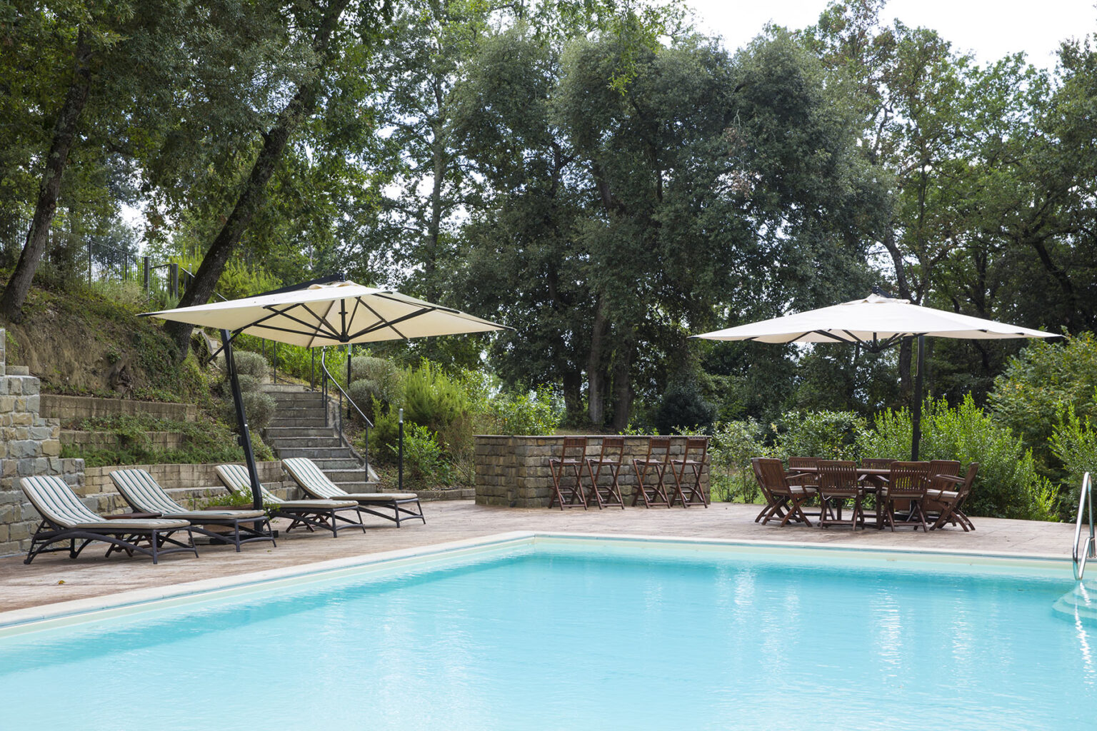 Serene outdoor swimming pool surrounded by lush greenery, featuring comfortable lounge chairs and large umbrellas for shade, ideal for relaxation and leisure. | Only on SopranoVillas