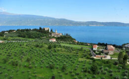 A panoramic view of Lake Garda, featuring lush olive groves, charming homes, and a historic castle on the hillside under a clear blue sky.