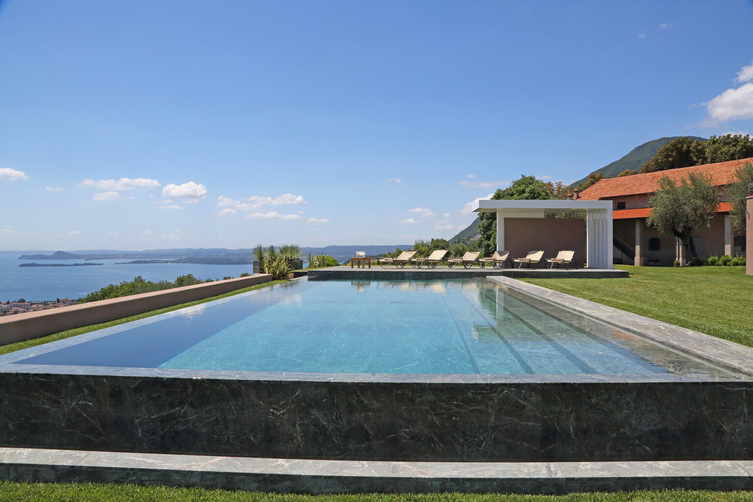 Luxury villa with an infinity pool overlooking a scenic lake and mountains, featuring a landscaped garden and sun loungers under a clear blue sky. | Only on SopranoVillas