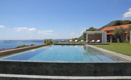 Luxury villa with an infinity pool overlooking a scenic lake and mountains, featuring a landscaped garden and sun loungers under a clear blue sky.