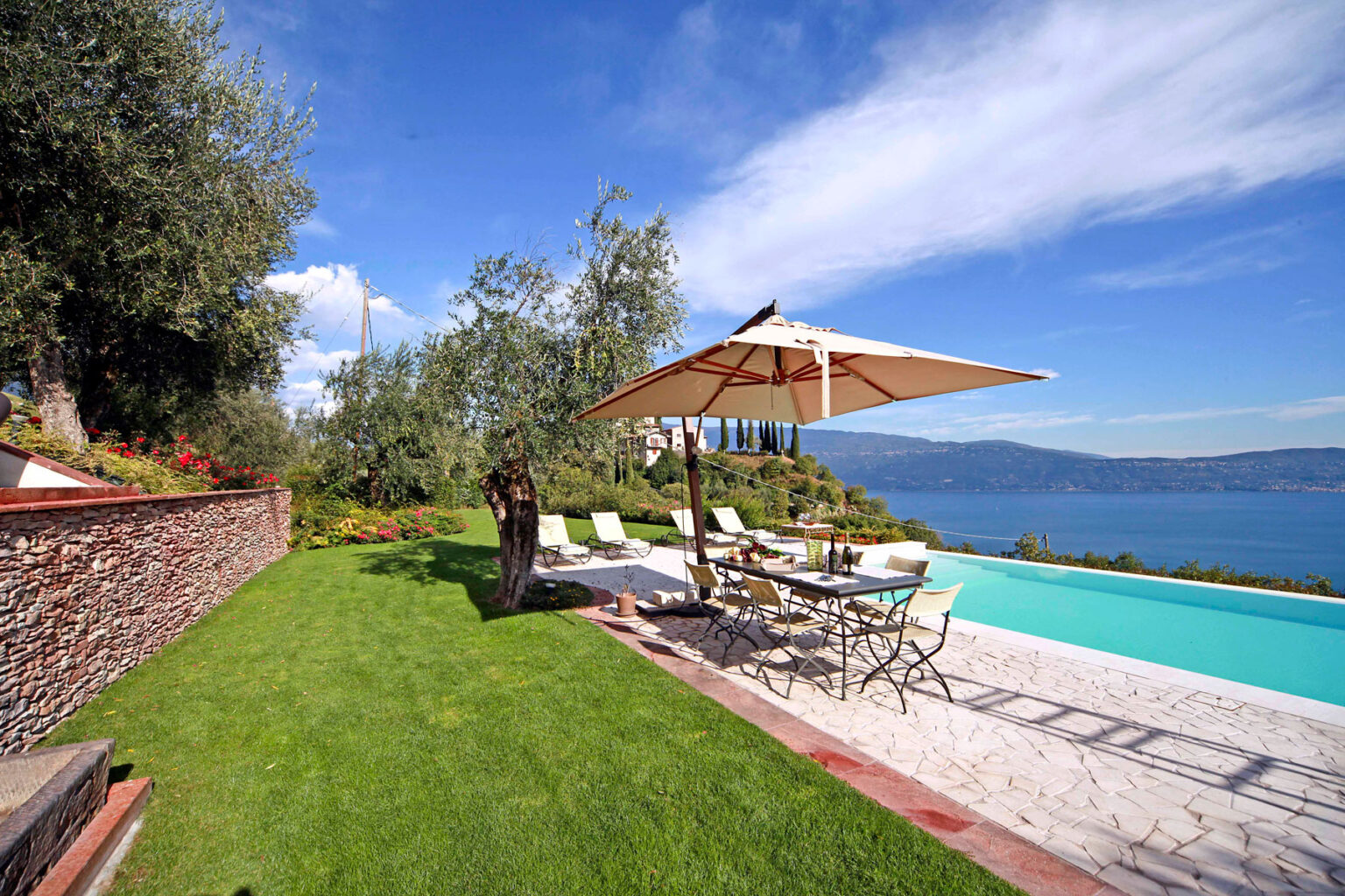 Beautiful outdoor pool area with sun loungers and an umbrella, overlooking a serene lake and lush greenery, ideal for relaxation and leisure activities. | Only on SopranoVillas