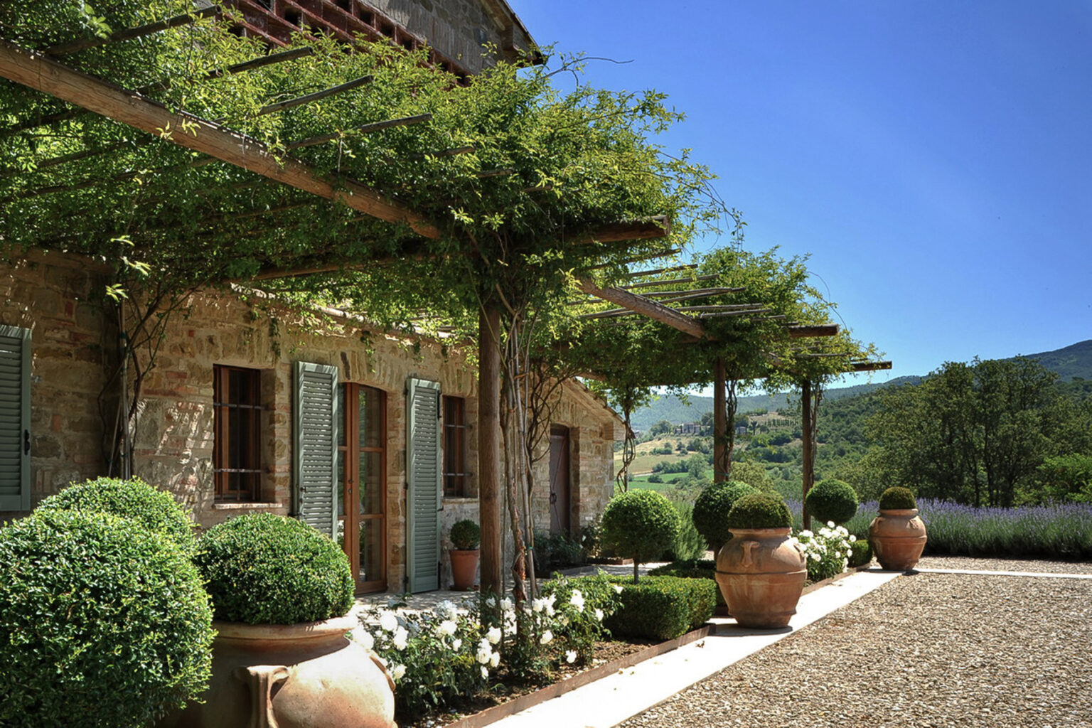 Idyllic Italian villa surrounded by lush greenery, featuring stone walls, wooden shutters, and decorative potted plants along a gravel pathway under a clear blue sky. | Only on SopranoVillas