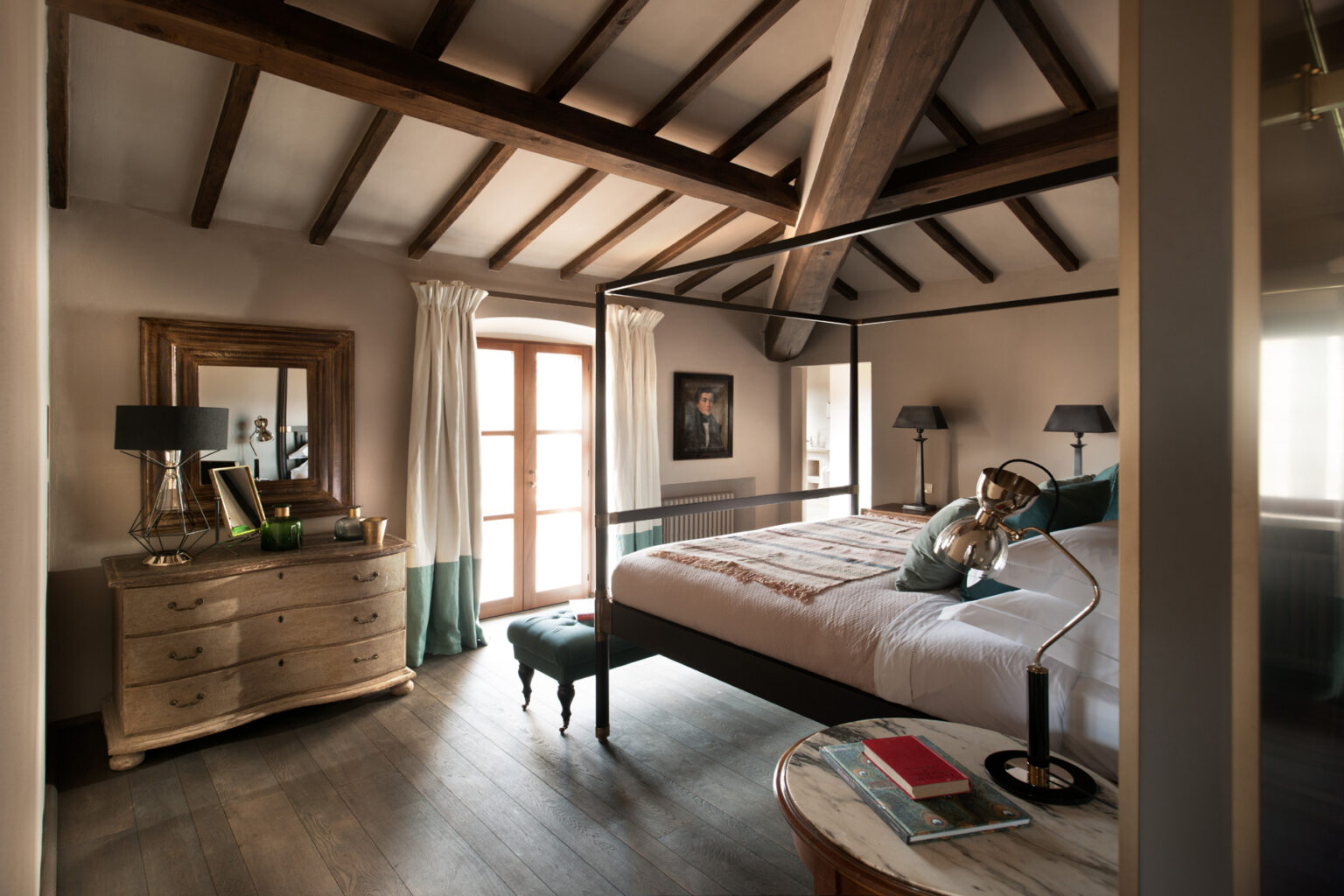 Cozy bedroom with a four-poster bed, wooden beams, and large windows. Features a stylish dresser, modern lamps, and decorative accents, creating a warm and inviting atmosphere. | Only on SopranoVillas