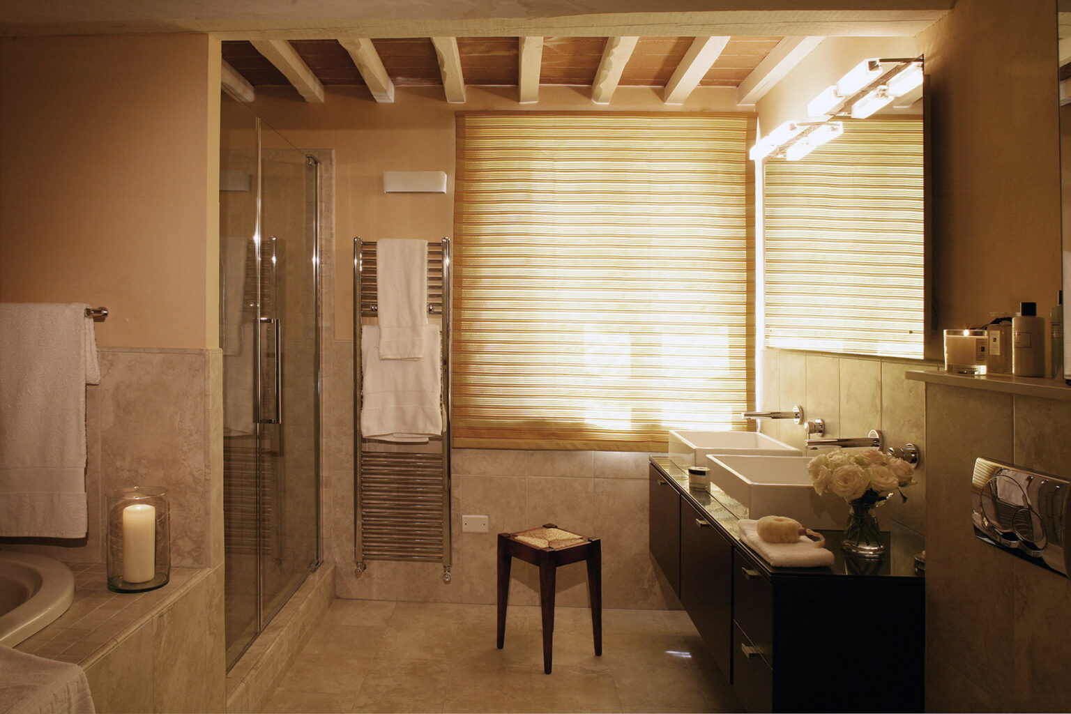 Modern bathroom interior featuring a glass shower, a double vanity with stylish fixtures, and natural light streaming through wooden blinds. The space is accented with neutral tones, a candle, and plush towels, creating a serene and inviting atmosphere. | Only on SopranoVillas