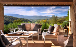 Cozy outdoor seating area with plush cushions overlooking a scenic mountain landscape, surrounded by greenery and bright blue skies, perfect for relaxation and enjoying nature.