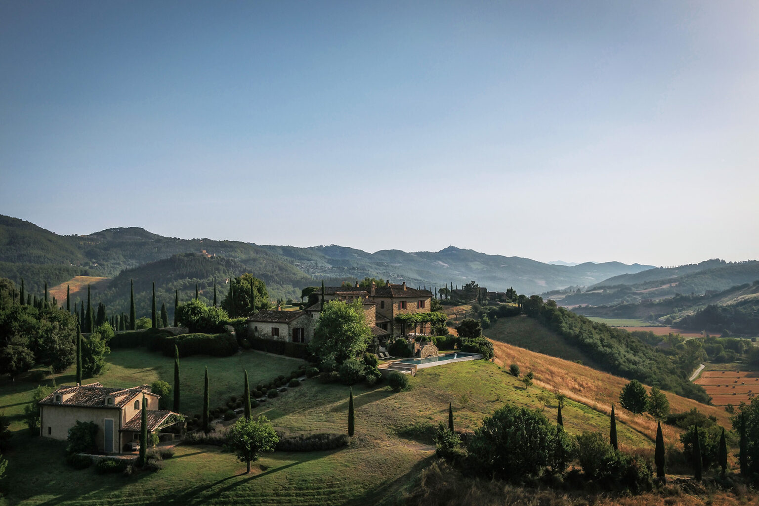Scenic view of a rustic villa in the Tuscan countryside, surrounded by rolling hills and vineyards under a clear blue sky. Ideal for travel enthusiasts and nature lovers. | Only on SopranoVillas