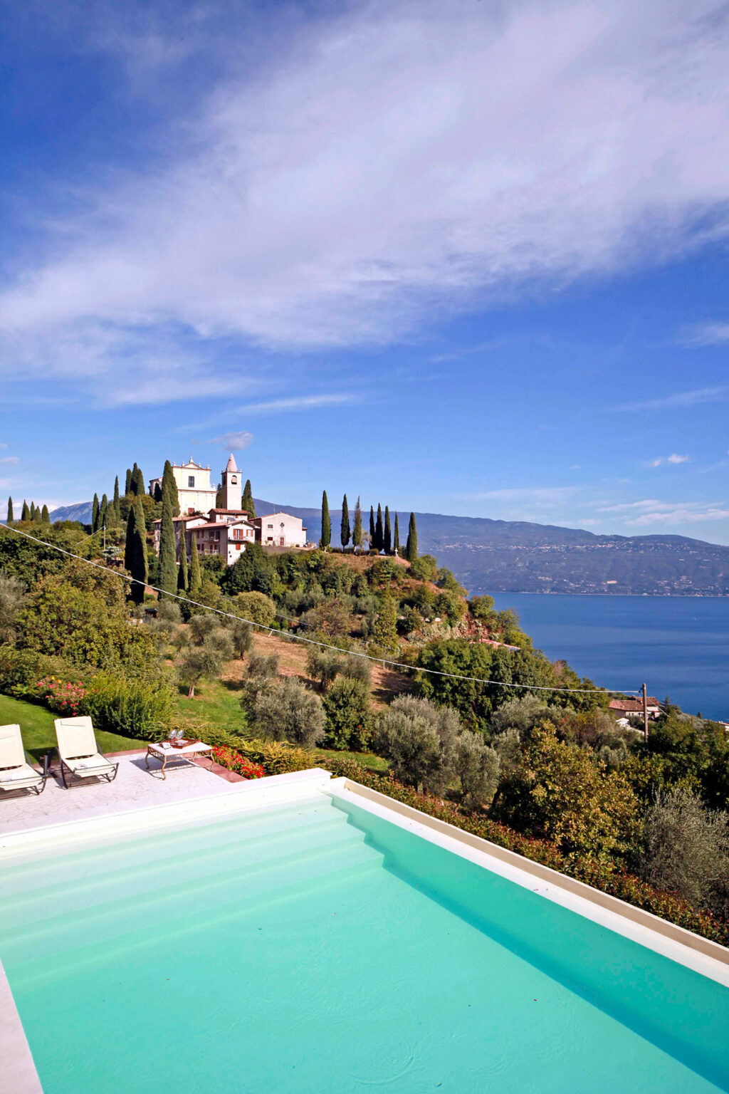 Scenic view of a hillside villa surrounded by cypress trees, overlooking a tranquil lake and mountains in the background, showcasing the beauty of Italian countryside architecture. | Only on SopranoVillas