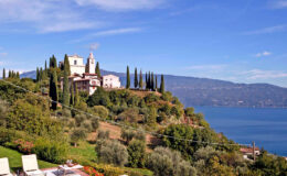 Scenic view of a hillside villa surrounded by cypress trees, overlooking a tranquil lake and mountains in the background, showcasing the beauty of Italian countryside architecture.