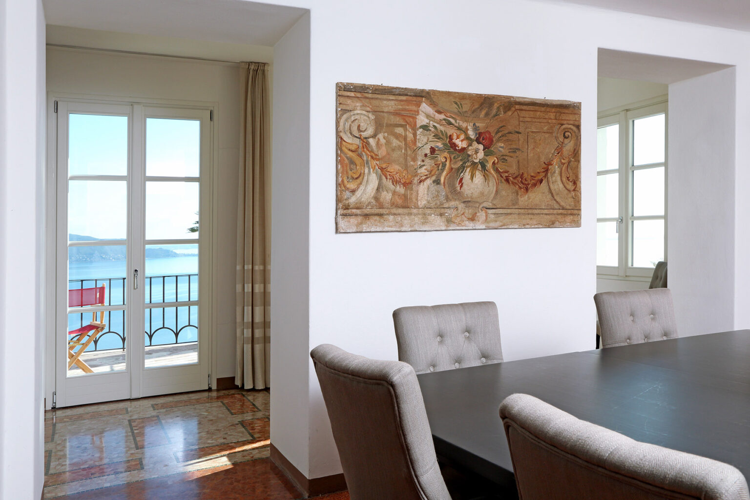 Modern dining room with a large black table and upholstered chairs, featuring a decorative wall mural and a view of the sea through glass doors. | Only on SopranoVillas