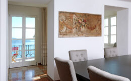 Modern dining room with a large black table and upholstered chairs, featuring a decorative wall mural and a view of the sea through glass doors.