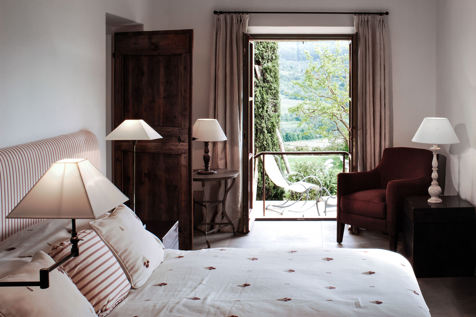 Cozy bedroom with a double bed, elegant lamps, and a view of a lush garden through open doors, featuring a comfortable armchair and tasteful decor. | Only on SopranoVillas