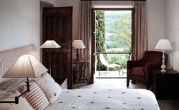 Cozy bedroom with a double bed, elegant lamps, and a view of a lush garden through open doors, featuring a comfortable armchair and tasteful decor.