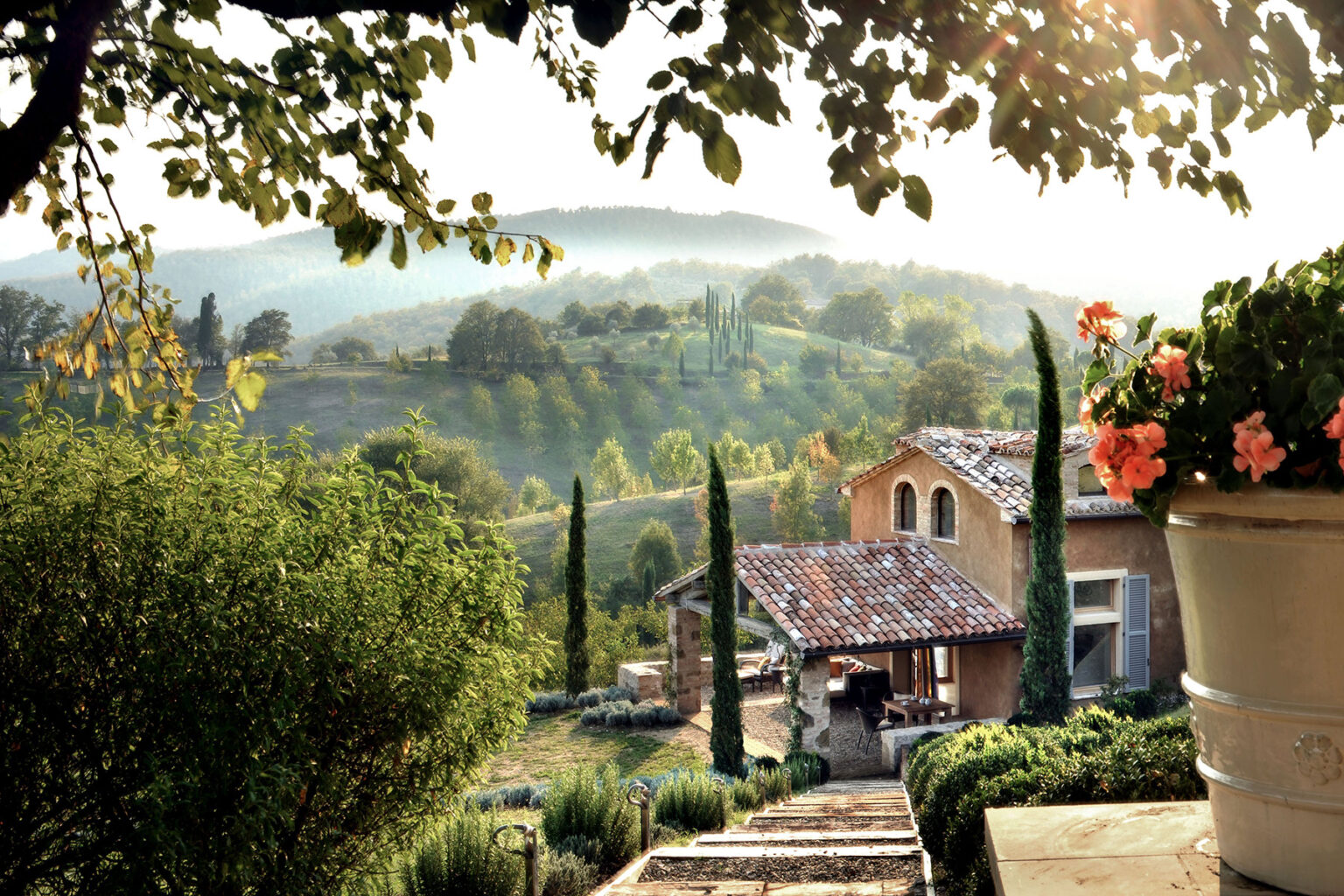 Scenic view of a rustic villa surrounded by lush greenery and cypress trees, with vibrant flowers in a pot, set against rolling hills and a misty morning sky. Ideal for promoting countryside retreats and nature tourism. | Only on SopranoVillas