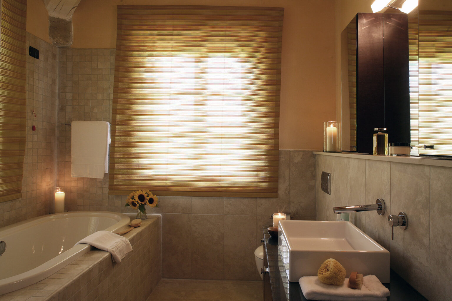 Modern bathroom interior featuring a soaking tub, a sleek sink, and natural light filtering through wooden blinds. Decorated with candles and a vase of sunflowers, the space exudes a warm and inviting atmosphere. | Only on SopranoVillas