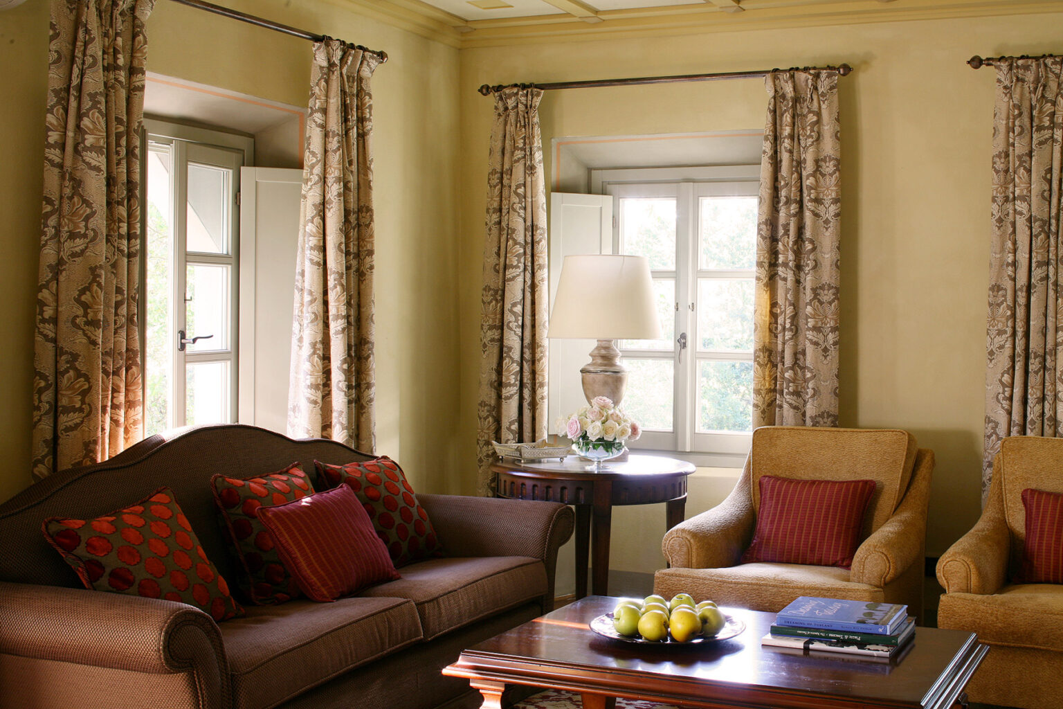 Cozy living room interior featuring a brown sofa with red accent pillows, two yellow armchairs, elegant curtains, and a wooden coffee table adorned with decorative items and fresh fruit. Natural light streams through the windows, enhancing the warm ambiance. | Only on SopranoVillas