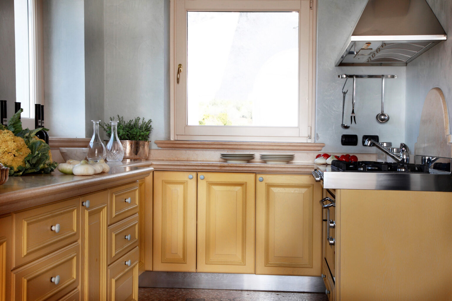 Modern kitchen featuring yellow cabinetry, a stainless steel countertop, and a window that brightens the space. The kitchen is equipped with a gas stove and hanging utensils, creating a functional and inviting cooking environment. | Only on SopranoVillas