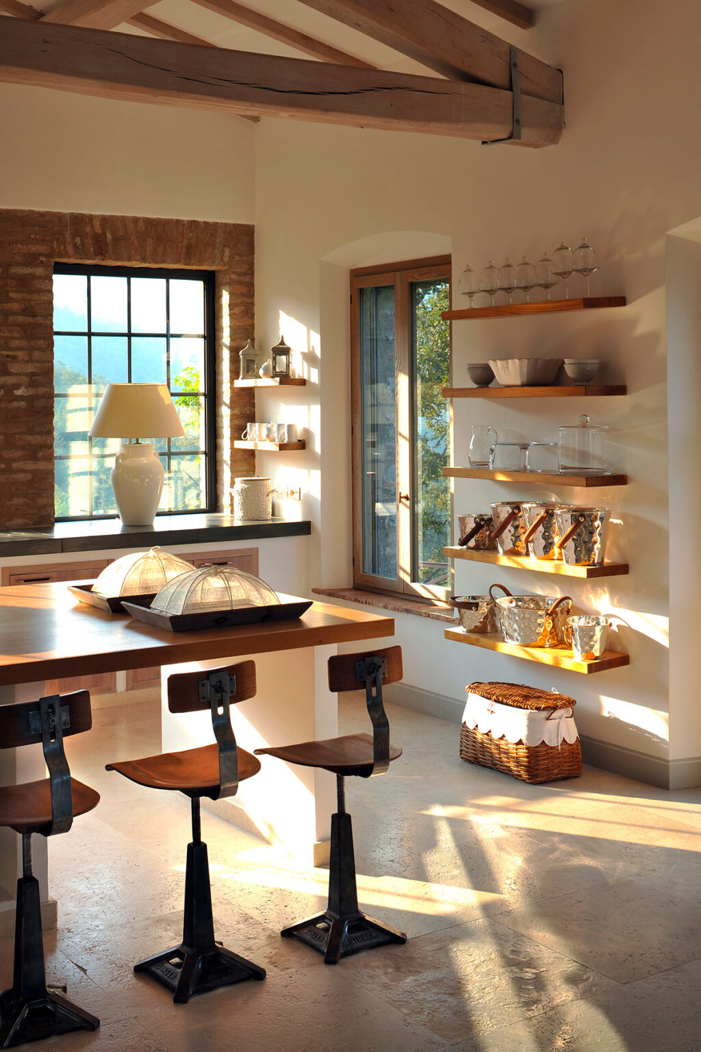 Bright and airy kitchen interior featuring a wooden dining table, stylish bar stools, and open shelves displaying elegant tableware. Natural light floods the space through large windows, highlighting the modern decor and warm color palette. | Only on SopranoVillas