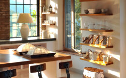 Bright and airy kitchen interior featuring a wooden dining table, stylish bar stools, and open shelves displaying elegant tableware. Natural light floods the space through large windows, highlighting the modern decor and warm color palette.