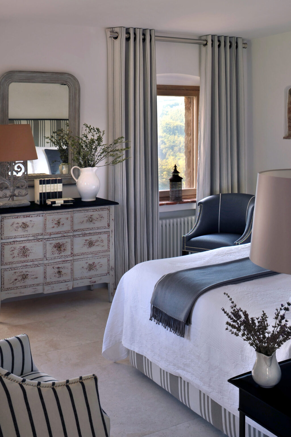 Elegant bedroom interior featuring a vintage dresser, a cozy armchair, and soft natural light streaming through a window. Decorative elements include a vase with flowers and stylish curtains, creating a serene and inviting atmosphere. | Only on SopranoVillas