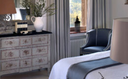 Elegant bedroom interior featuring a vintage dresser, a cozy armchair, and soft natural light streaming through a window. Decorative elements include a vase with flowers and stylish curtains, creating a serene and inviting atmosphere.