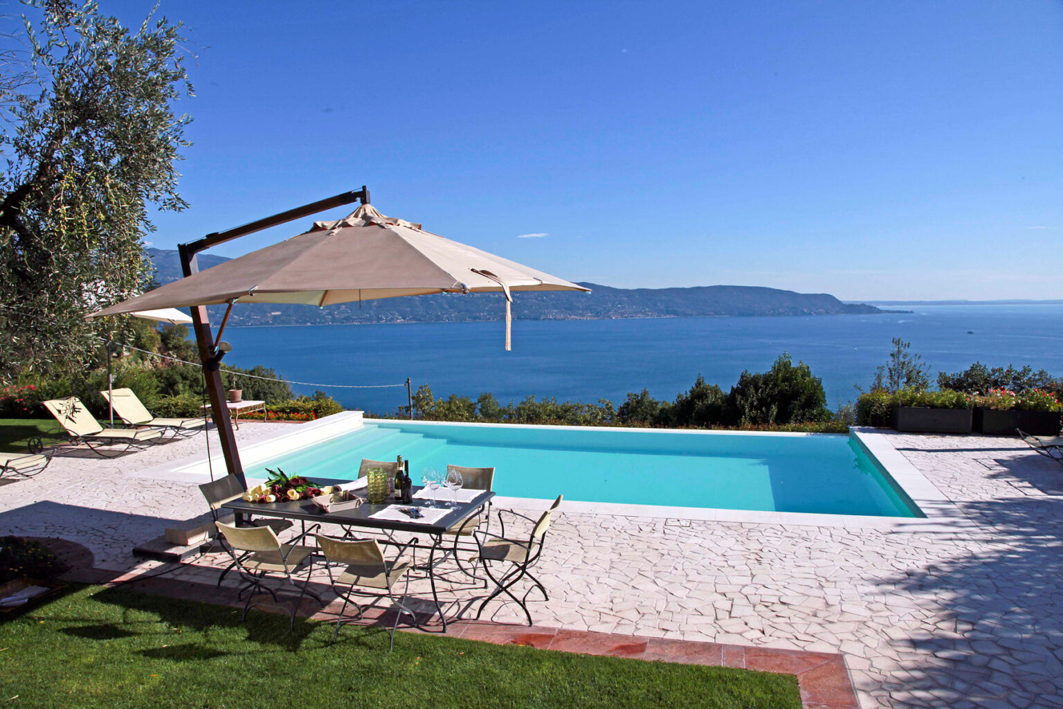 Luxury villa with a swimming pool overlooking a serene lake, featuring sun loungers, a dining area under a large umbrella, and lush greenery in the background. Ideal for relaxation and outdoor gatherings. | Only on SopranoVillas