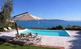 Luxury villa with a swimming pool overlooking a serene lake, featuring sun loungers, a dining area under a large umbrella, and lush greenery in the background. Ideal for relaxation and outdoor gatherings.