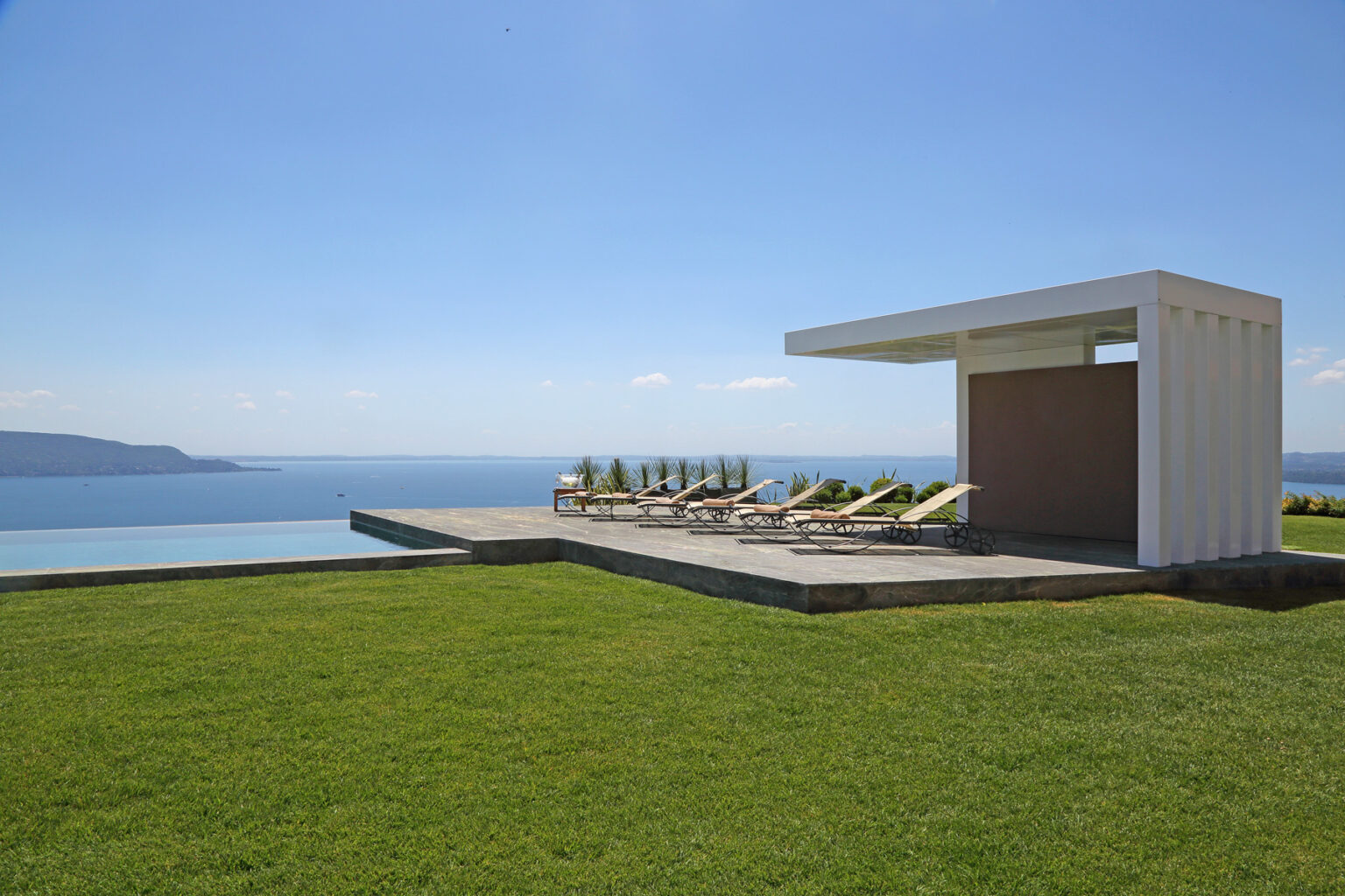 Modern outdoor lounge area with sun loungers overlooking a serene ocean view, featuring a sleek white cabana and lush green grass under a clear blue sky. Ideal for relaxation and leisure. | Only on SopranoVillas