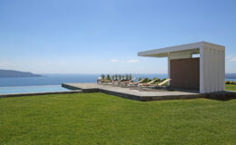 Modern outdoor lounge area with sun loungers overlooking a serene ocean view, featuring a sleek white cabana and lush green grass under a clear blue sky. Ideal for relaxation and leisure.