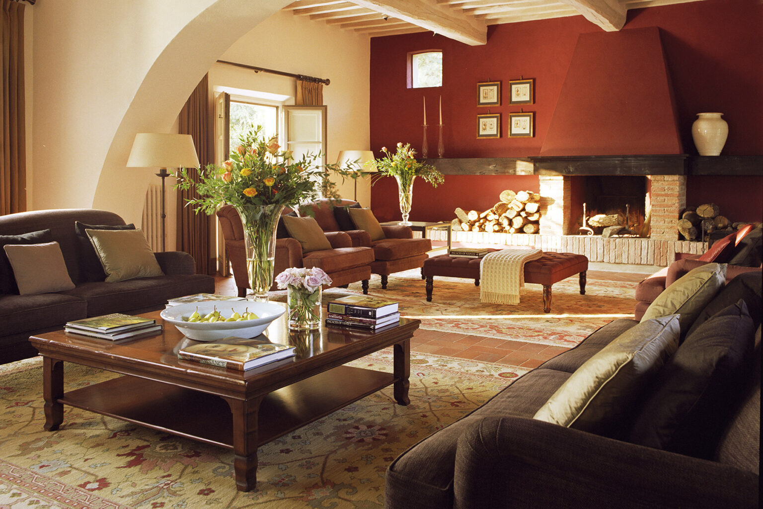 Cozy living room with a red accent wall, featuring stylish brown sofas, a wooden coffee table with books and a decorative bowl, and a fireplace surrounded by logs. Brightened by natural light from large windows and adorned with fresh flowers, the space exudes warmth and comfort. | Only on SopranoVillas