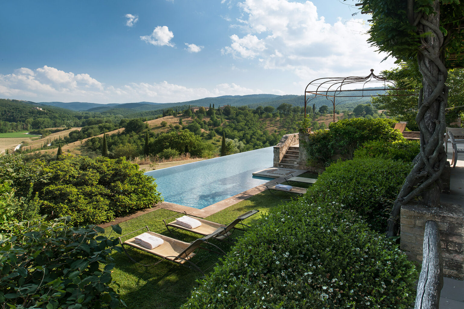 Scenic view of a luxury villa with an infinity pool overlooking rolling green hills and a blue sky, featuring sun loungers and landscaped gardens. | Only on SopranoVillas